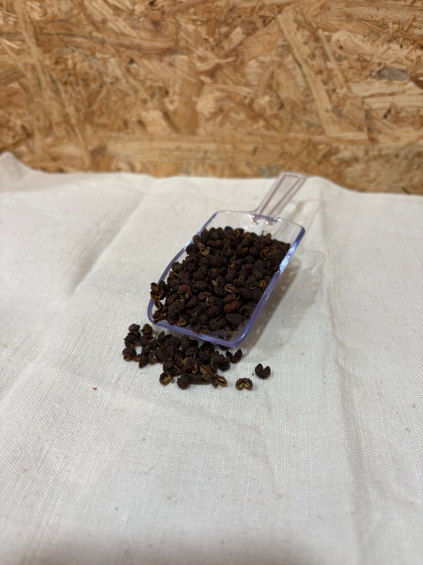 Pimenta Sichuan Pá transparente com grãos pequenos escuros sobre tecido branco e fundo de madeira aparente