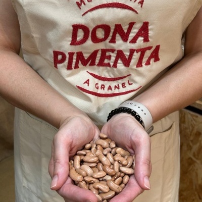 Mãos segurando feijão castanho com avental bordado MERCEARIA DONA PIMENTA A GRANEL