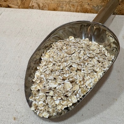 Pá metálica com flocos de aveia crus e cabo de madeira sobre tecido branco e fundo de madeira OSB