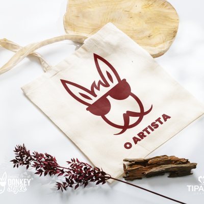 Saco de pano com desenho bordô de rosto de animal e texto O ARTISTA sobre fundo branco com madeira e ramo seco