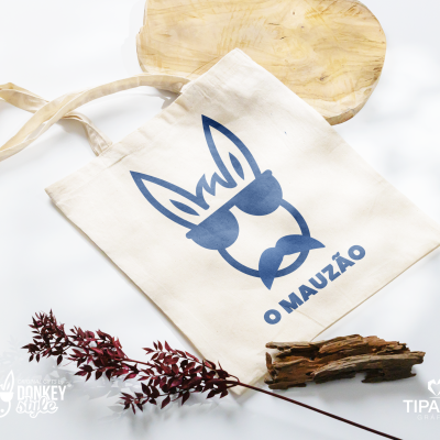 Saco de tecido creme com impressão azul de rosto estilizado e texto O MAUZÃO
