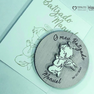 Produto redondo em madeira com anjo gravado e texto do batizado sobre embalagem branca com ilustração e texto dourado