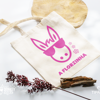 Saco de tecido branco com estampa rosa de um burro de óculos de sol e texto 'A FLORZINHA', rodeado por elementos naturais