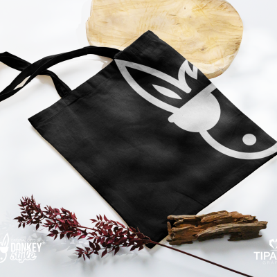 Mala tote preta em tecido com desenho branco de burro junto a ramos secos vermelhos e pedaços de madeira sobre base de madeira clara