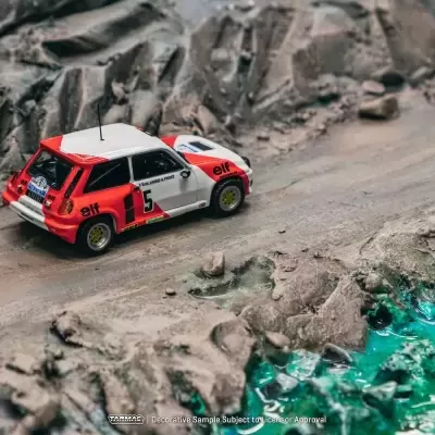 Miniatura de carro de rali vermelho e branco, número 5, em estrada de terra com pedras e água azul.
