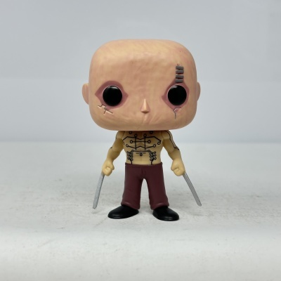 Figura Funko Pop com cabeça grande e cicatrizes, corpo tatuado, calças castanhas e duas espadas cinzentas.