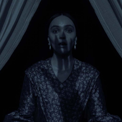 Mulher de vestido floral escuro e brincos pendentes, entre cortinas brancas