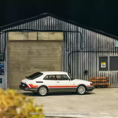 Miniatura de carro Saab branco com faixas vermelhas e laranja junto a edifício industrial em metal ondulado