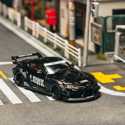 Miniatura de carro desportivo preto Liberty Walk numa pista com linhas brancas e amarelas, junto a vedação e postes.