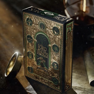 Caixa de baralho de cartas Lord of the Rings em verde escuro e dourado com símbolos e texto, ao lado de uma aliança dourada e rolos de pergaminho