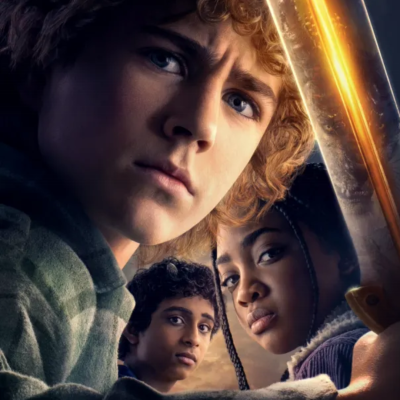 Três jovens com expressões sérias, um loiro com camisola verde, uma rapariga com objeto iluminado, e um rapaz com camisola castanha