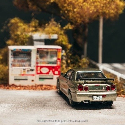 Modelo reduzido de carro desportivo verde metálico em cenário de rua com máquinas de venda automática e árvores amarelas