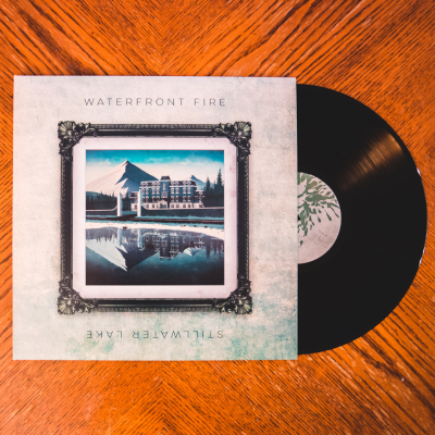 Disco de vinil preto com capa ilustrada de hotel junto a lago e montanhas.