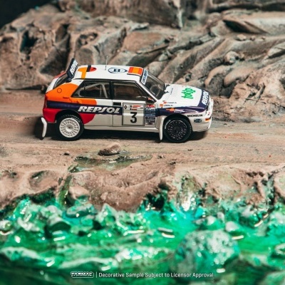 Miniatura de carro de rali branco com logótipos coloridos numa estrada de terra junto a água verde