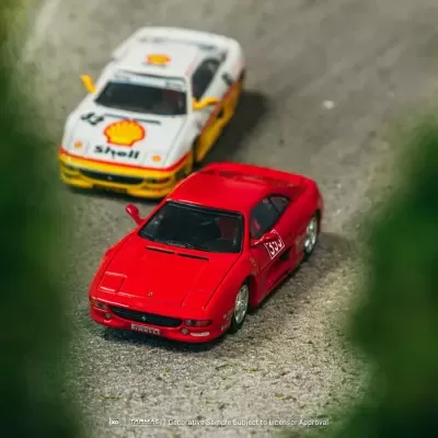 Dois carros de brinquedo em miniatura, um vermelho e um branco com logo Shell, sobre superfície cinzenta rugosa