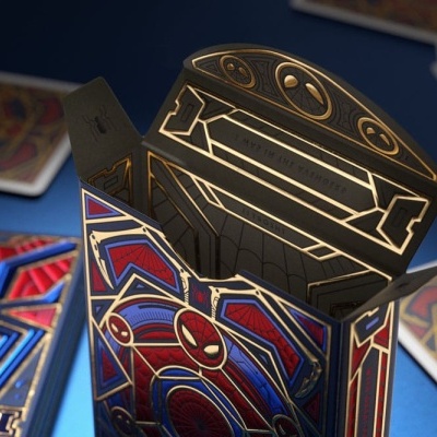 Caixa metálica decorativa Homem-Aranha com embalagem interna preta e dourada sobre mesa azul com cartas de baralho