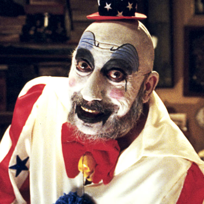 Palhaço com maquilhagem branca e azul, chapéu preto com estrelas e gravata borboleta vermelha.