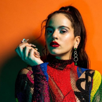 Mulher com blusa colorida brilhante, brincos de círculos e anéis, fundo laranja.