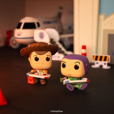 Duas figuras Funko Pop de Toy Story com roupas distintas, uma de cowboy e outra de astronauta, segurando caixas vermelhas, com avião branco ao fundo.