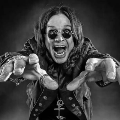 Homem de cabelo comprido com óculos e casaco escuro apontando mãos para a frente.