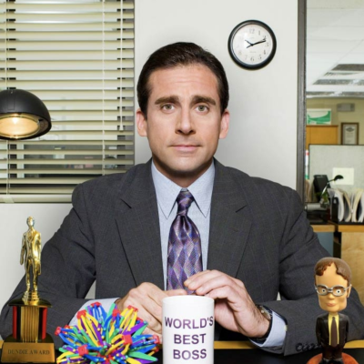 Homem vestido formal sentado à secretária com caneca WORLD'S BEST BOSS e troféu Dundie Award