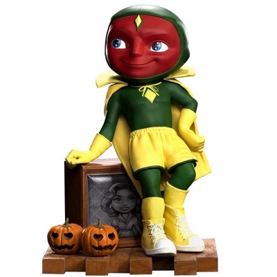 Estatueta de super-herói com cabeça vermelha em forma de maçã, fatos verde e amarelo, duas abóboras de Halloween e placa desenhada