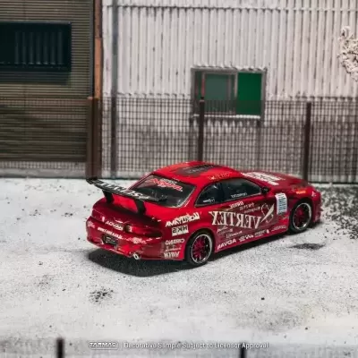 Carro miniatura de corrida vermelho com textos e logos brancos e pretos, com spoiler e rodas vermelhas, estacionado junto a cerca metálica e edifício.