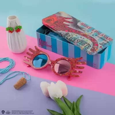 Óculos de sol com armação em formato de mão em rosa e vermelho, caixa azul às riscas, brincos de fruta vermelha e verde e flores brancas sobre fundo rosa e lilás.