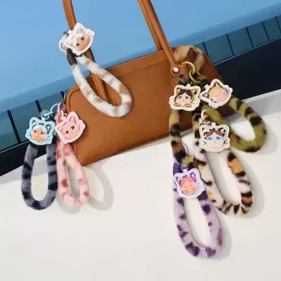 Porta-chaves e pulseiras de pelo com desenho de rosto de gato pendurados numa mala castanha sobre superfície branca