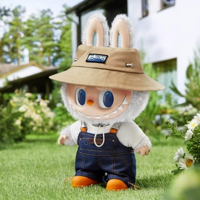 Boneco de peluche com macacão, chapéu bege THE MONSTERS, olhos azuis grandes, na relva com flores