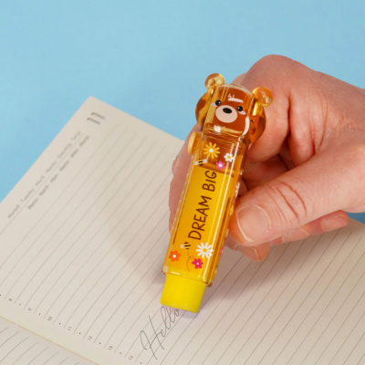 Corrector amarelo em forma de urso com flores e frase DREAM BIG, sendo usado numa agenda aberta.