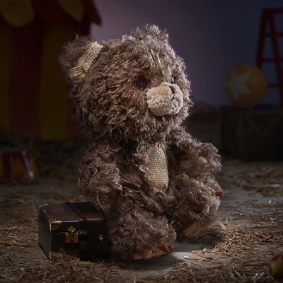 Urso de pelúcia castanho com detalhes em juta a sentar junto a uma mala num chão com palha