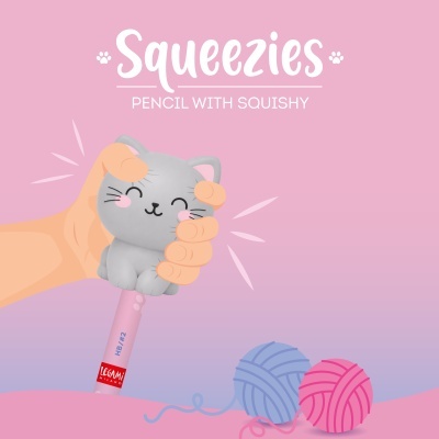 Lápis com topo de gato cinzento squishy sendo apertado por uma mão, lápis rosa com texto HB/#2 e bolas de lã azul e rosa ao lado