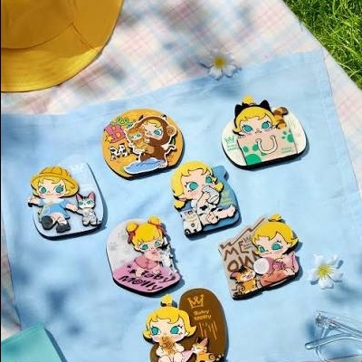 Sete pins Baby Molly Baby Tabby Series dispostos em pano azul claro sobre manta xadrez pastel com relva e chapéu amarelo ao lado.