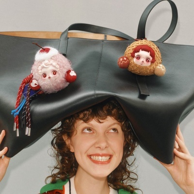 Mala de mão preta com pendurados decorativos de bonecas coloridas segurada por mulher sorridente