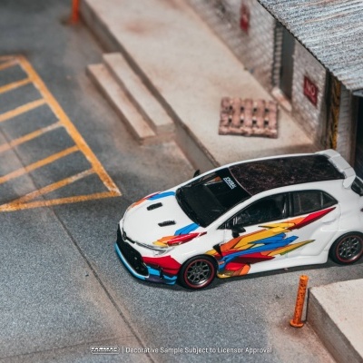 Carro de brinquedo branco com gráficos coloridos numa miniatura de rua e porta de garagem azul