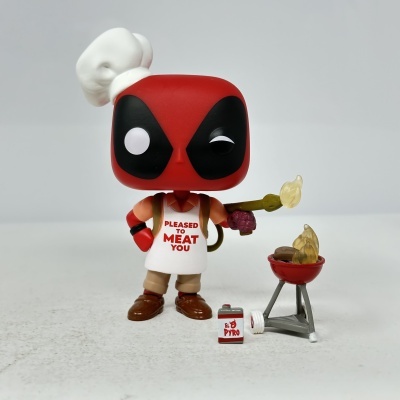 Figura Funko Pop Deadpool com avental branco e lança-chamas ao lado de churrasqueira vermelha