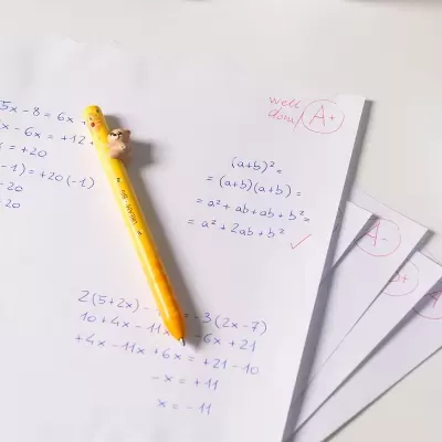 Folhas com equações matemáticas e correção, caneta amarela com ursinho sobre fundo branco.