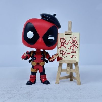 Figura Funko Pop do Deadpool pintor junto a quadro em cavalete com desenhos vermelhos