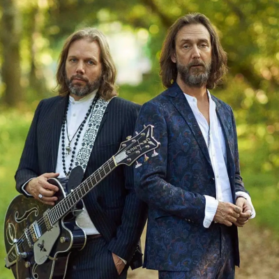 Dois homens de fato, um com guitarra elétrica, em ambiente natural com árvores atrás.