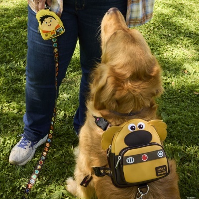 Mochila para cão castanha com design de rosto de cão colocada nas costas de um golden retriever, junto a pessoa com calças jeans e trela colorida.