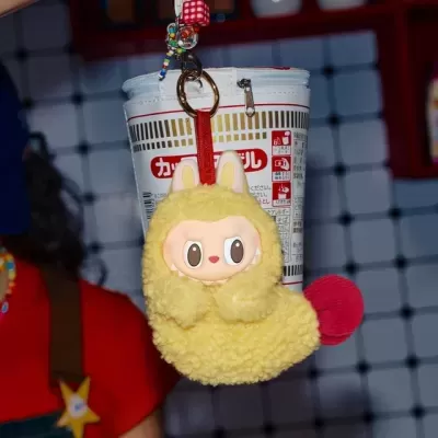 Chaveiro de pelúcia amarelo com cabeça de boneco e cauda vermelha pendurado em embalagem de plástico