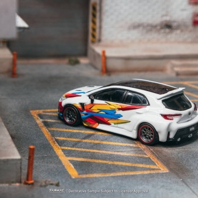 Carro em miniatura branco com padrão geométrico colorido e jantes pretas com aro vermelho num cenário urbano.