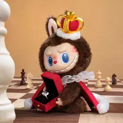 Boneco de peluche com coroa e caixa vermelha com anel em tabuleiro de xadrez.
