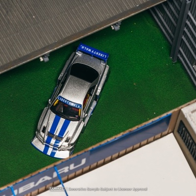 Miniatura de carro desportivo prateado com riscas azuis e texto LIBERTY WALK em cenário de parque com relva artificial.
