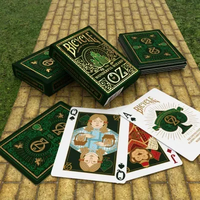 Baralhos de cartas temáticos The Wonderful Wizard of Oz com caixas e cartas exibindo design verde e dourado colocados sobre um chão de tijolos amarelos ao ar livre.