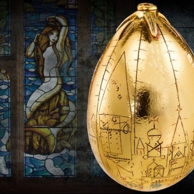 Ovo dourado decorativo com desenhos de castelos à frente de vitral com figura feminina