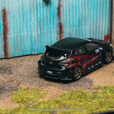 Carro em miniatura preto com gráficos vermelhos e roxos em chão de terra perto de portão azul enferrujado