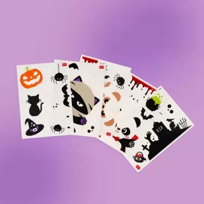 Folhas com autocolantes de Halloween, incluindo abóbora, gato preto, aranha, múmia e panda, sobre fundo roxo.
