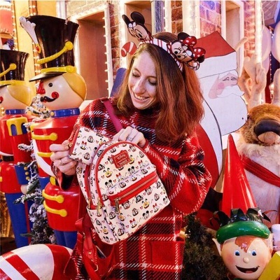 Mochila pequena de tecido bege com estampado de personagens Disney e zíperes vermelhos, mulher com suéter vermelho xadrez numa decoração natalícia.